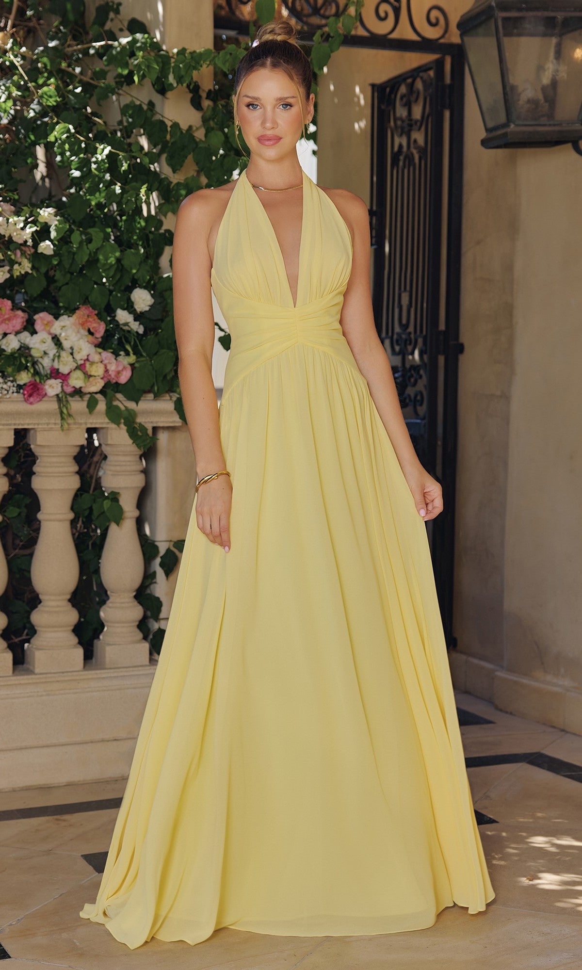 A woman stands indoors wearing the Ladivine T715, a light yellow, sleeveless, floor-length chiffon halter prom dress. Marble flooring, floral arrangement, and wrought iron gate appear in the background.